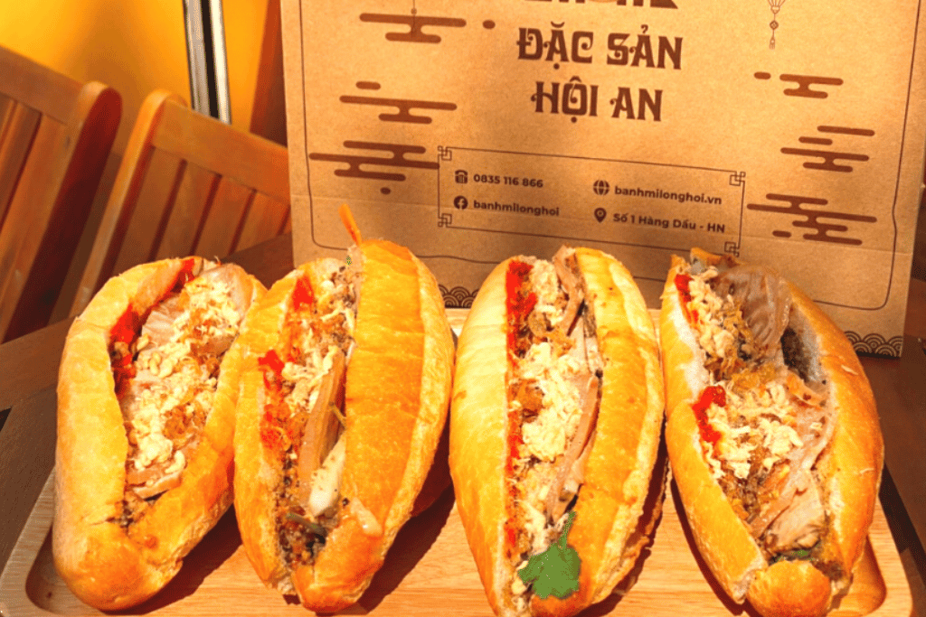 Four filled banh mi baguettes on a wooden board beside a Banh Mi Long Hoi paper bag