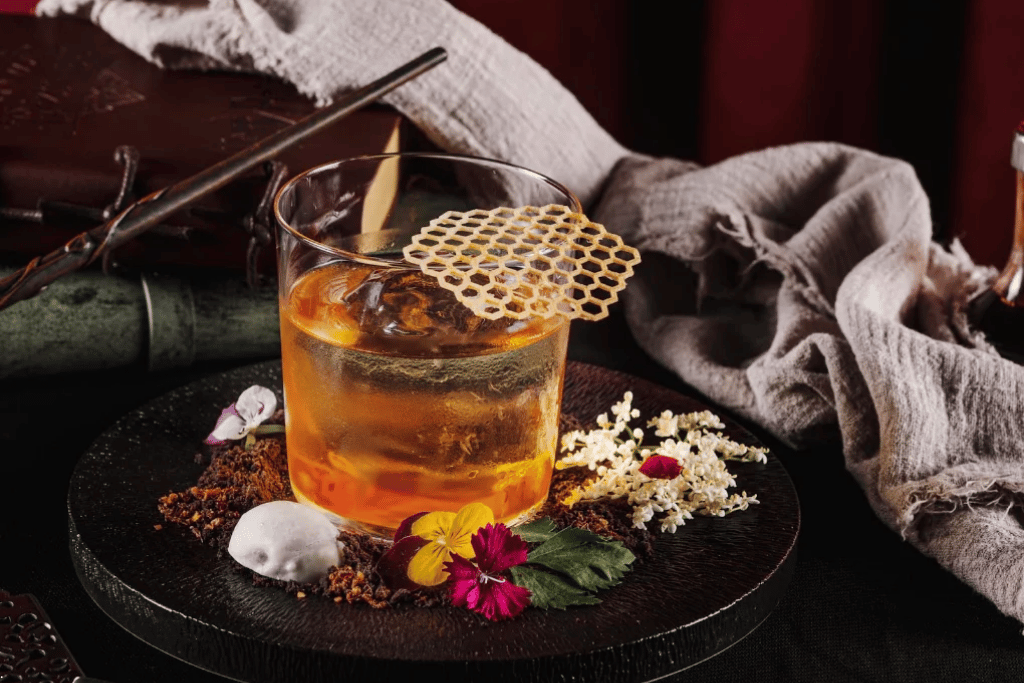 Amber cocktail garnished with a honeycomb tuile and edible flowers on a dark slate board