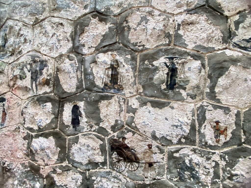 Small painted figures of conical-hat vendors and a rickshaw on a textured stone mural wall