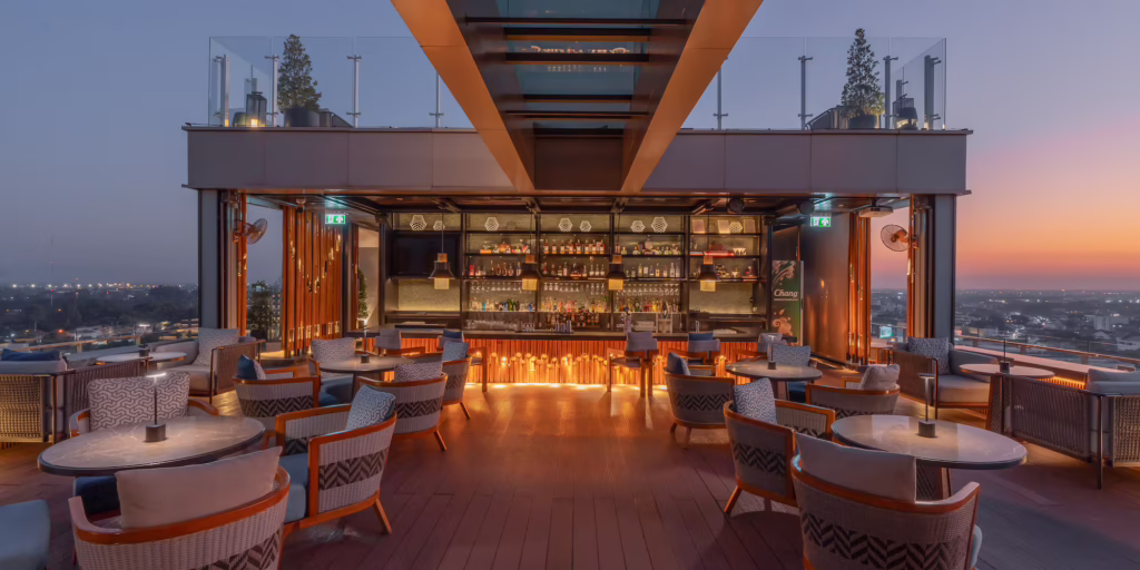 An open-air rooftop bar with lounge seating and a backlit bar overlooking the city at sunset