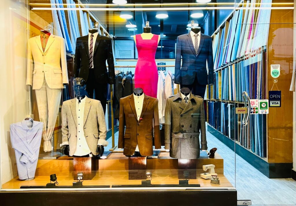 Mannequins in bespoke suits and a pink dress in a Chiang Mai tailor's window, fabric bolts behind