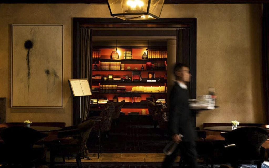 A dimly lit bistro interior with a lounge-style book nook and a waiter carrying drinks