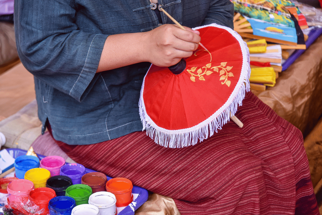 An artisan hand-painting a gold floral design onto a red paper umbrella, paint pots in front