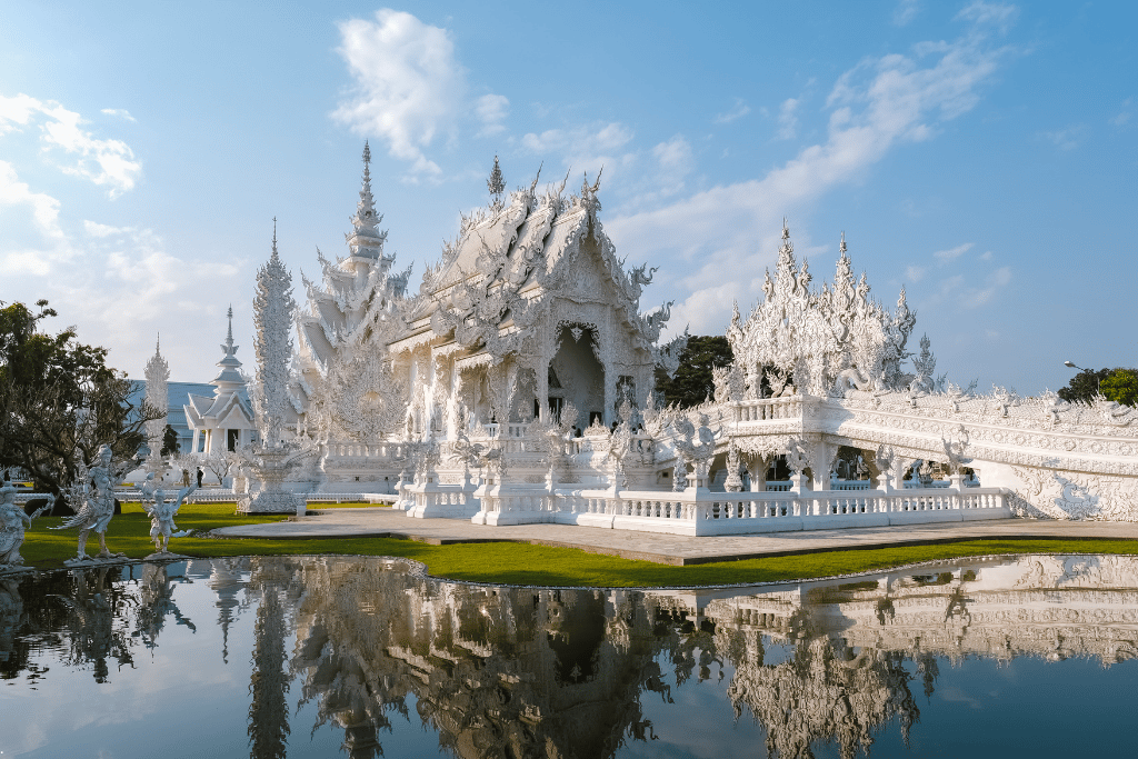The ornate all-white Wat Rong Khun temple mirrored in a pond under a blue sky in Chiang Rai