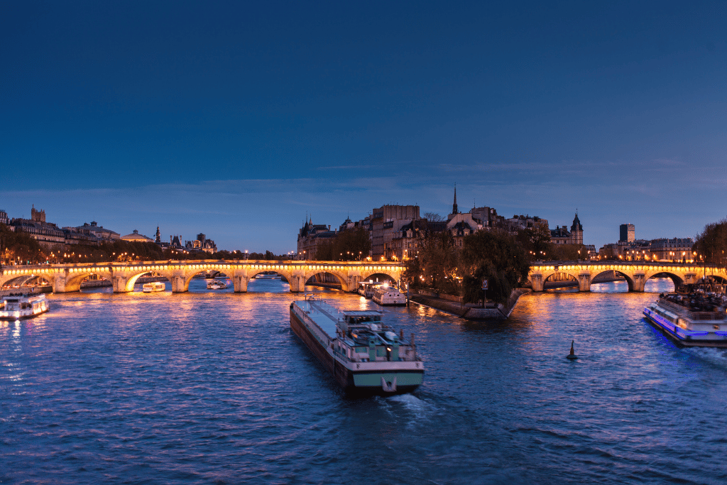 A river barge passing under a lit bridge on the Seine near Île de la Cité at dusk