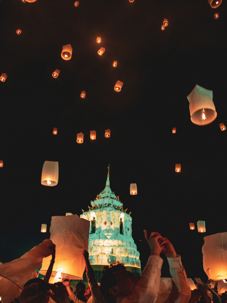 Paper sky lanterns drifting above an illuminated temple spire at Chiang Mai's Yi Peng festival
