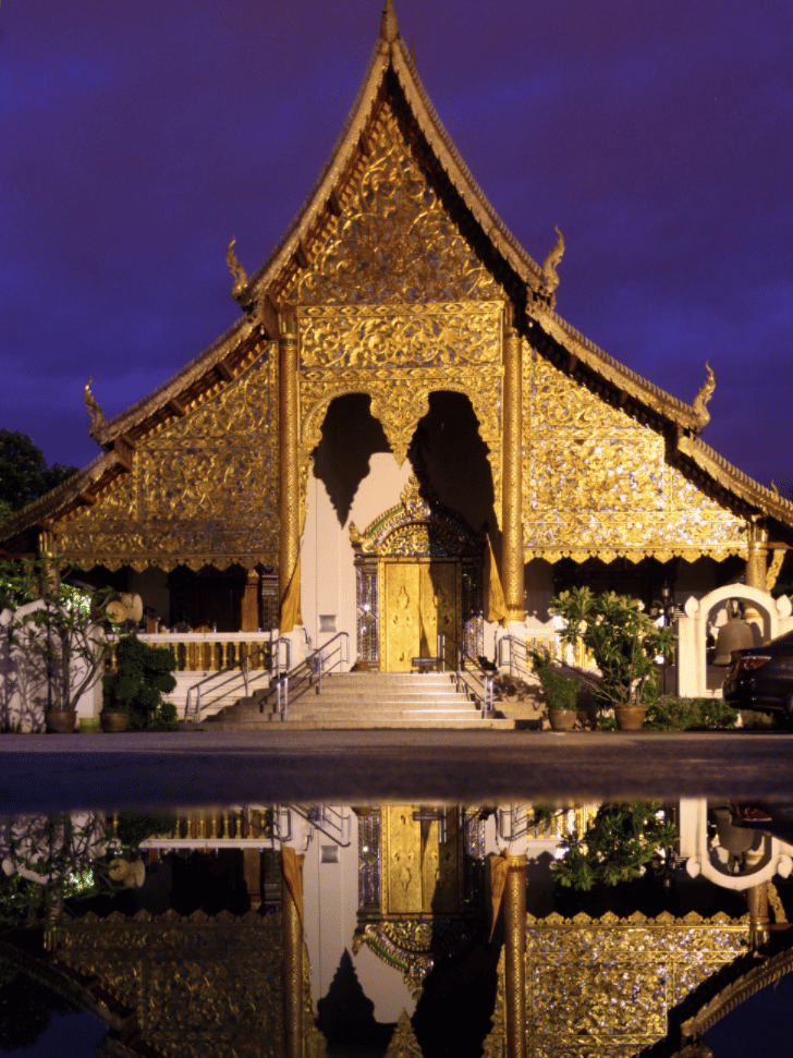 An ornate gold-and-teak Lanna temple hall mirrored in still water at dusk
