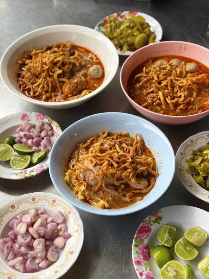 Three bowls of Khao Soi topped with crispy noodles, with lime, shallots and pickled greens alongside