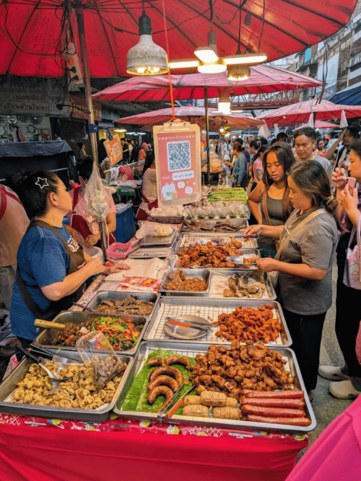 Trays of fried snacks and sausages under red umbrellas at a busy Warorot Market food stall