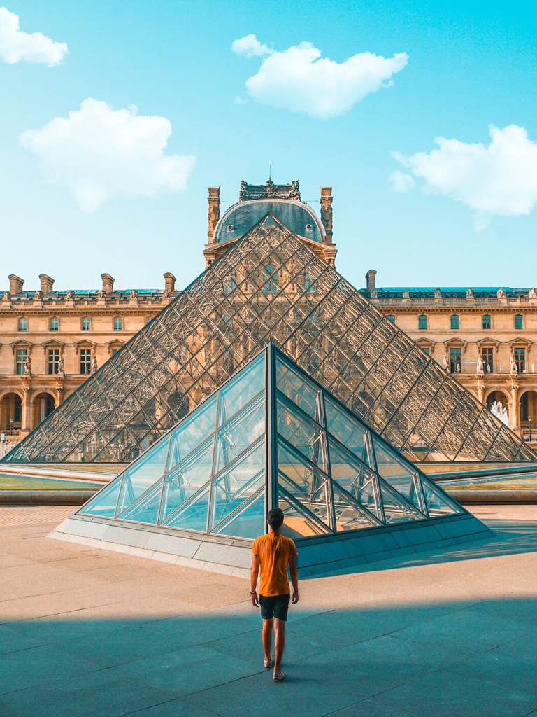 Visitor walking toward the Louvre's glass pyramid entrance with the museum building behind