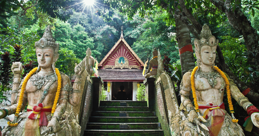Guardian statues flanking a mossy staircase up to Wat Pha Lat's shrine in the forest