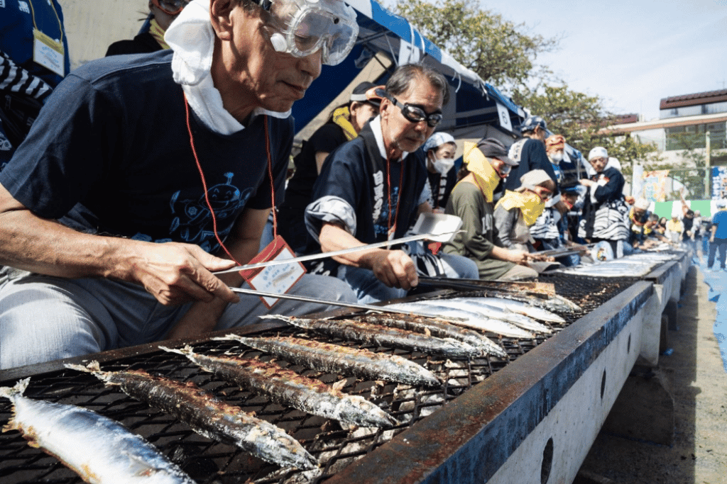 People grilling rows of pacific saury fish over a long charcoal grill at a festival