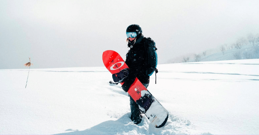 Snowboarder carrying a red board walking through deep fresh powder snow on a slope