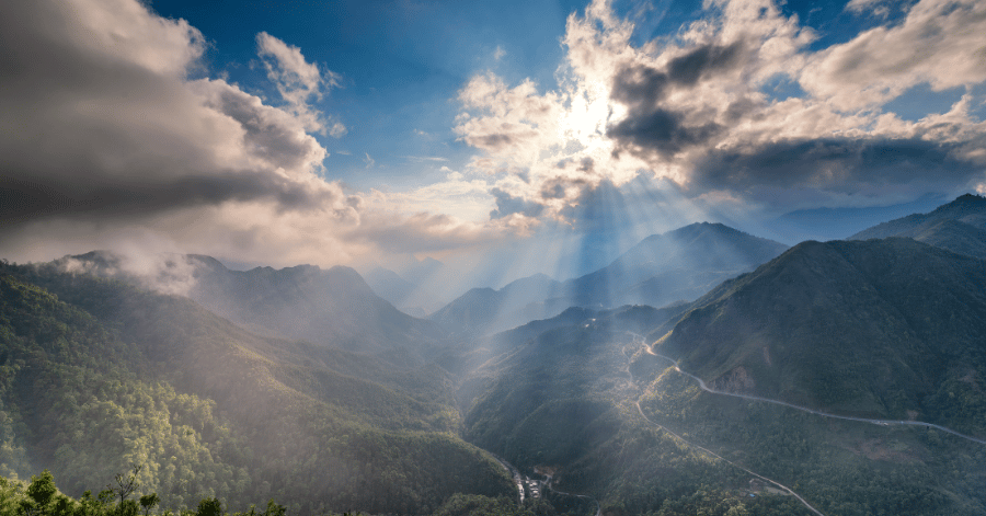 Sun rays breaking through clouds over a forested mountain valley and winding road near Sapa