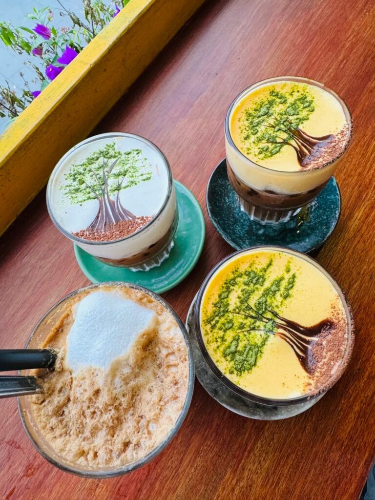 Four Vietnamese coffees topped with green tree-shaped latte art on a wooden table by a window