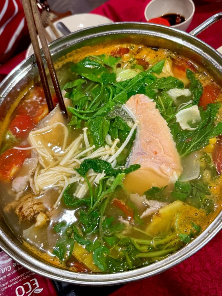 A steaming salmon hot pot with herbs, tomatoes and enoki mushrooms in a metal pot