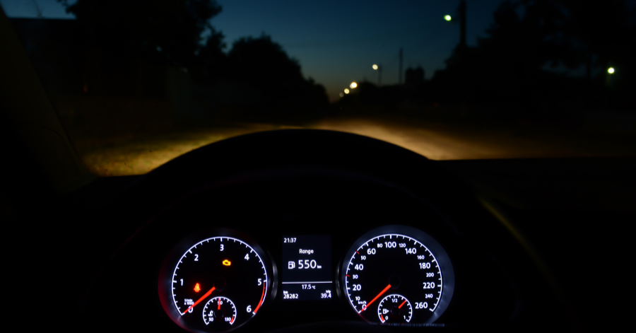 View from inside a car at night, lit dashboard and headlights on a dark road