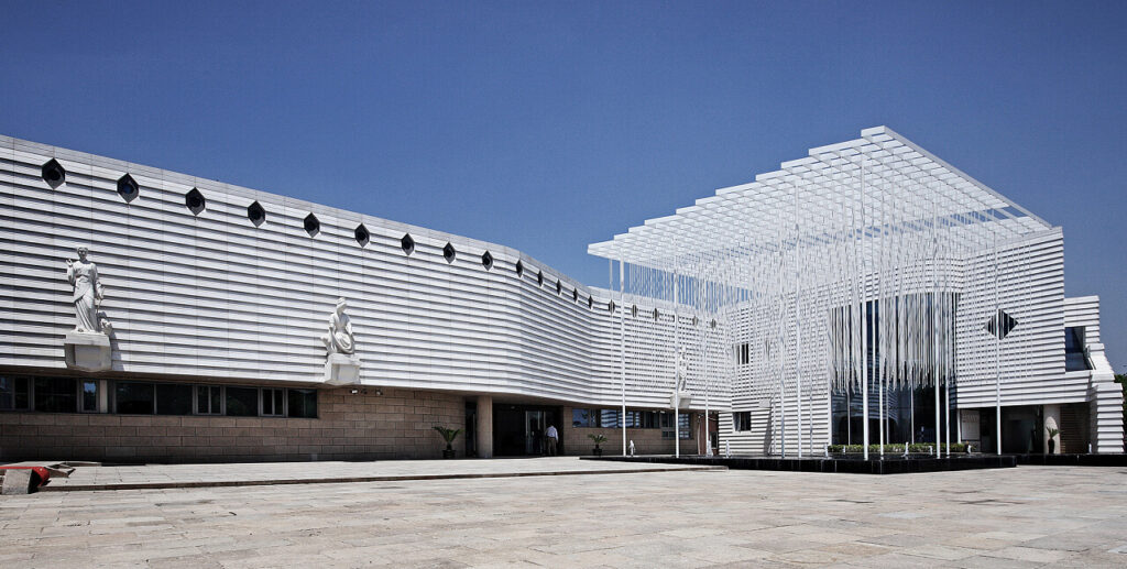 A modern white louvred building with classical statues and an open lattice pavilion