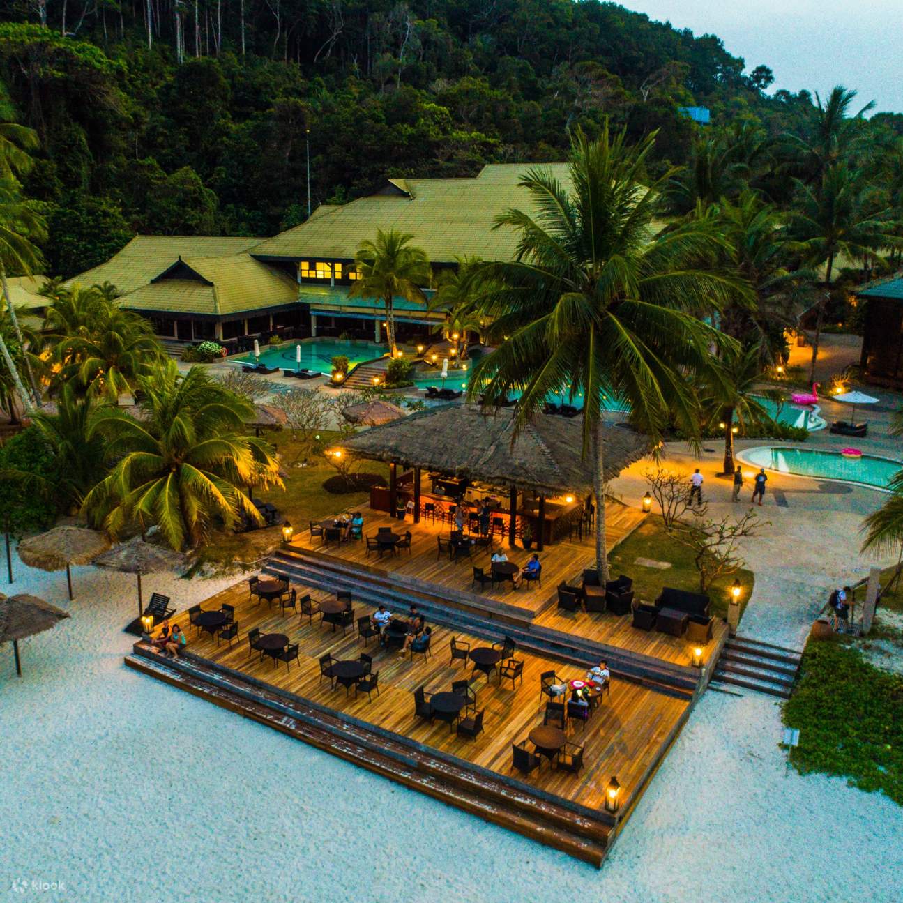 Aerial view of a beachfront resort with a lit wooden dining deck, pools and palms at dusk