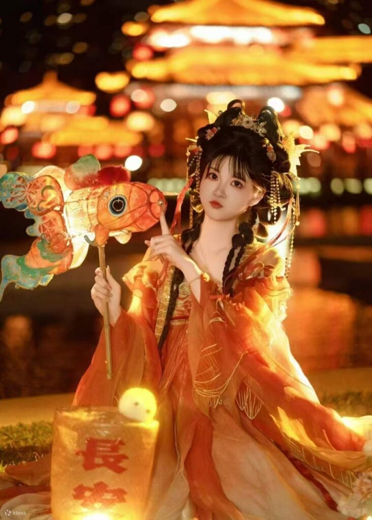 A woman in flowing orange hanfu holds a fish lantern at night in Xi'an