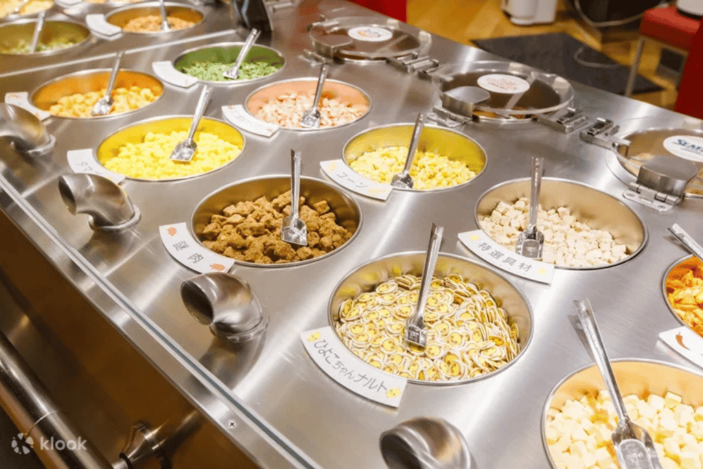 Stainless steel toppings bar with a dozen cup noodle ingredient choices and scoops