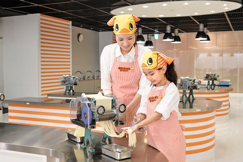 Parent and child in chicken hats and aprons making noodles at the Chicken Ramen Factory