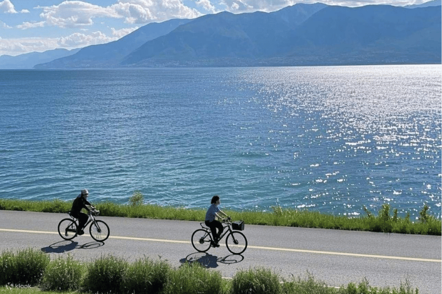 Two cyclists riding a lakeside road beside Erhai Lake with mountains behind, Dali