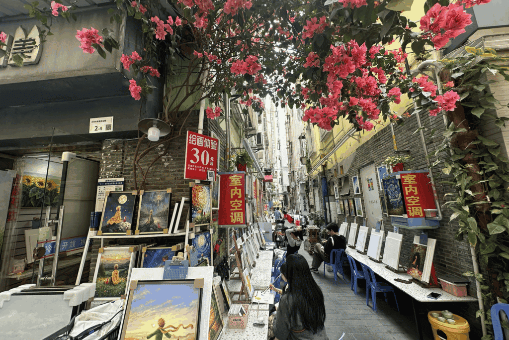 Paintings displayed along a narrow alley hung with red flowers at Dafen art village