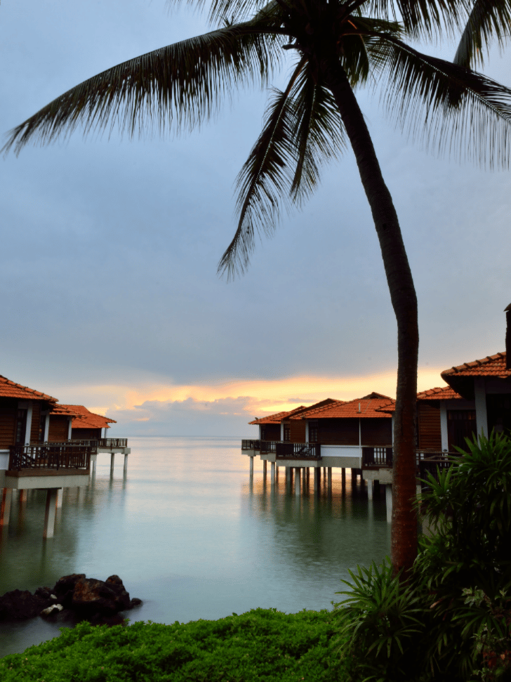 Over-water villas framed by a palm tree at dusk in Port Dickson