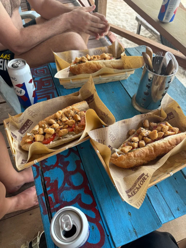 Three prawn-filled baguette sandwiches in paper trays on a blue table with drinks