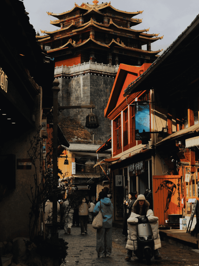 People walking a Yunnan old-town lane below a tall traditional timber tower