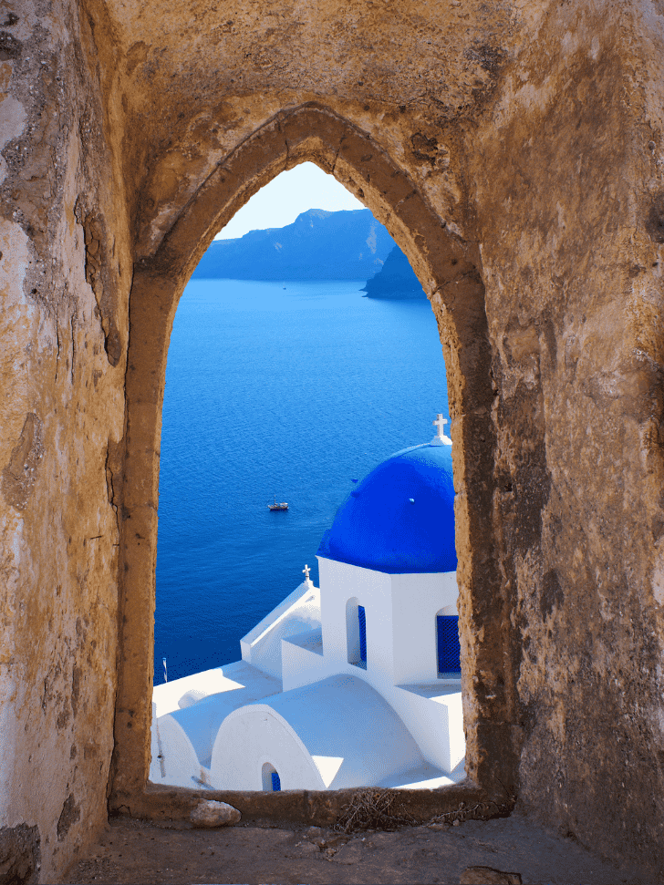 A blue-domed Santorini church framed through an old stone archway above the blue sea
