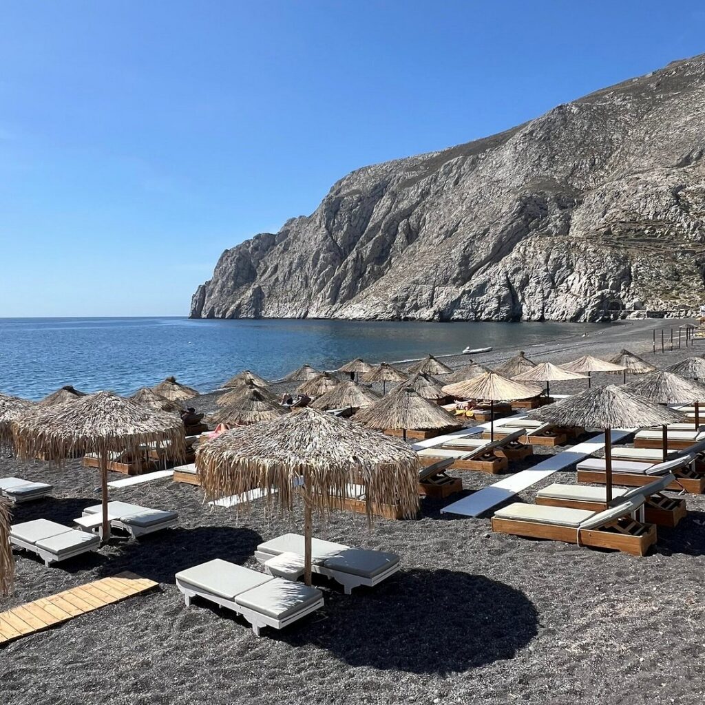 Straw umbrellas and sunbeds on a black-sand beach below tall cliffs, Santorini
