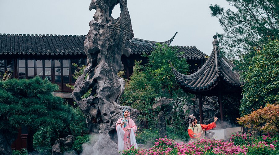 Two performers in traditional dress beside a towering scholar's rock in a Suzhou garden