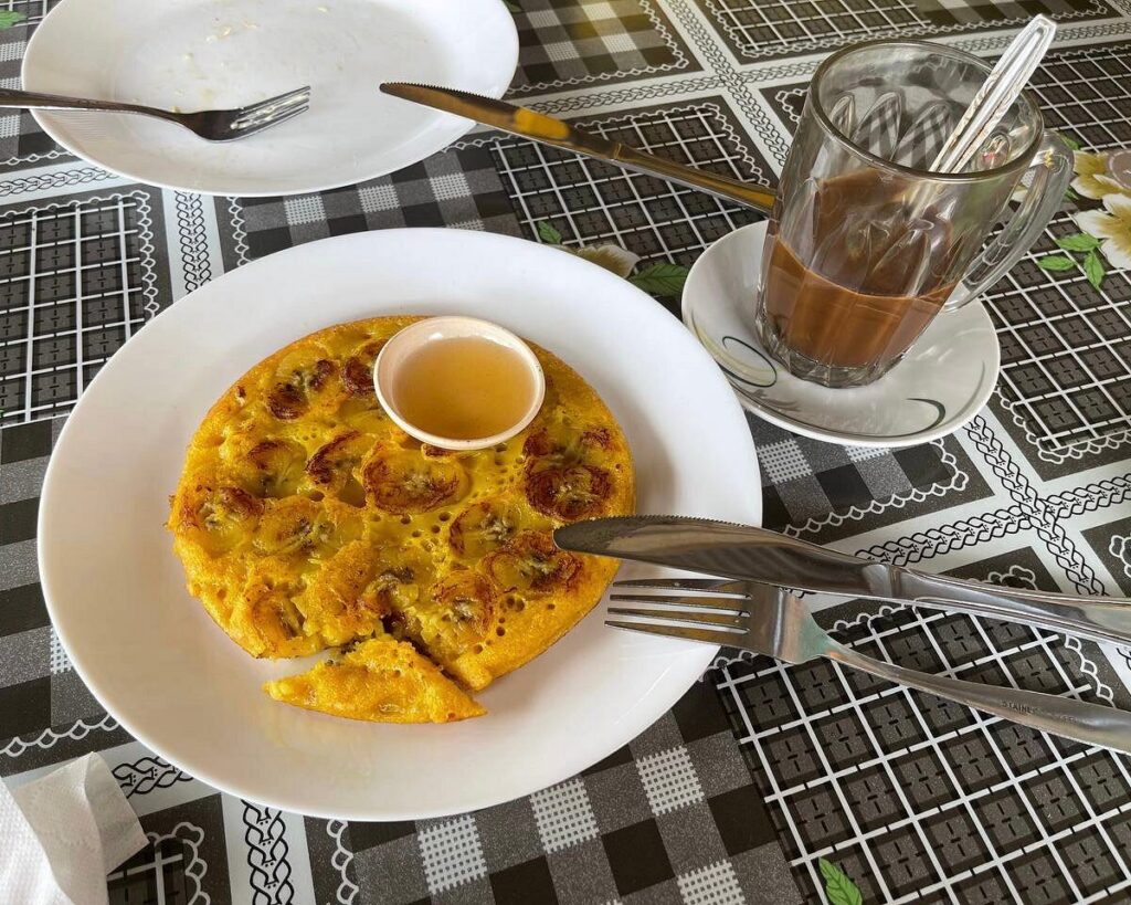 A plate of banana pancake with honey beside a cup of tea on a cafe table