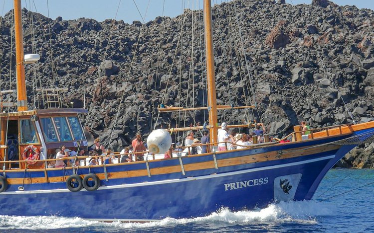 A wooden tour boat full of passengers passing the black volcanic rock of Nea Kameni