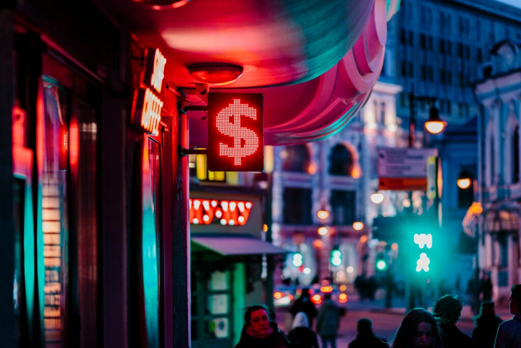 A glowing red dollar-sign currency exchange sign on a busy city street at dusk
