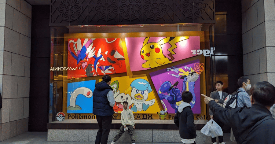 Street-level storefront window with colourful Pokémon artwork panels as pedestrians walk past in Shinsaibashi, Osaka