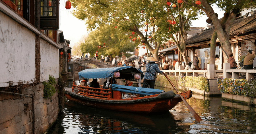 A boatman punting a traditional wooden boat along a tree-lined Suzhou canal