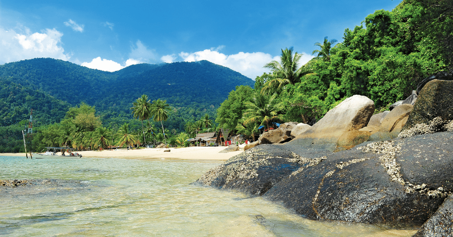 Granite boulders on a Tioman beach beside clear shallow water and forested hills