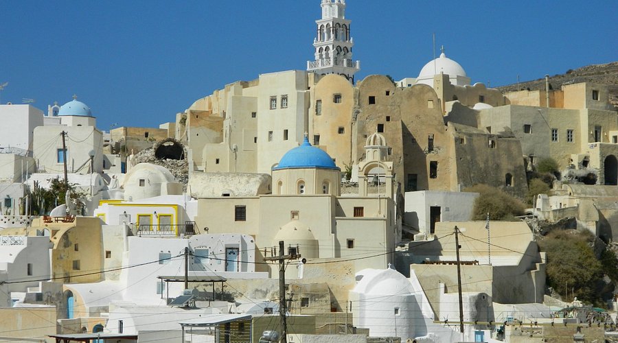 A hillside Santorini village of cream and white houses with a blue dome and tall bell tower