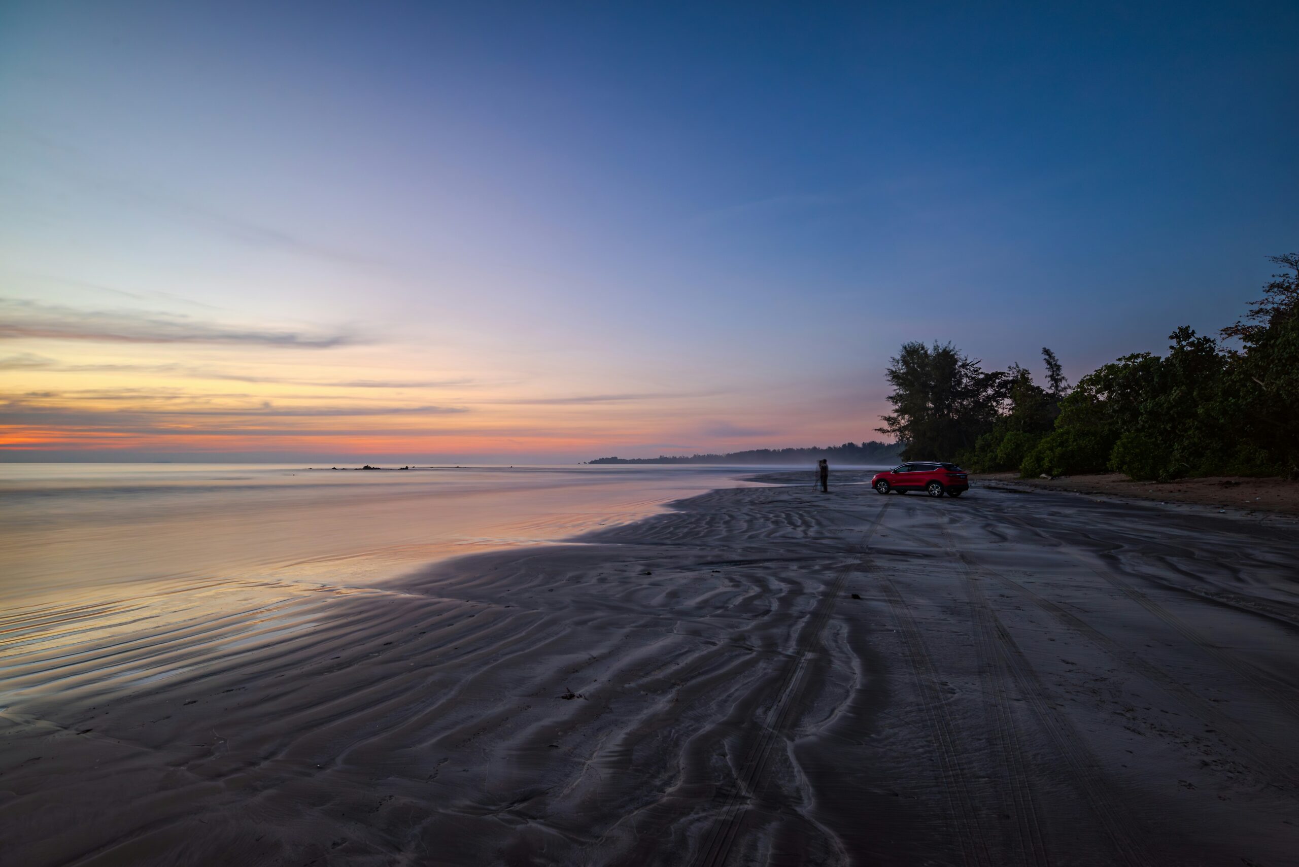 A red SUV parked on Desaru's wet-sand beach as the sun sets over a calm sea