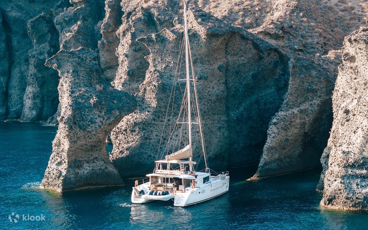 A white sailing catamaran anchored in clear water beside pale Santorini sea cliffs
