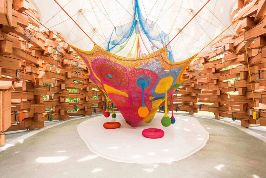 A large colourful crocheted net play sculpture inside a wooden dome