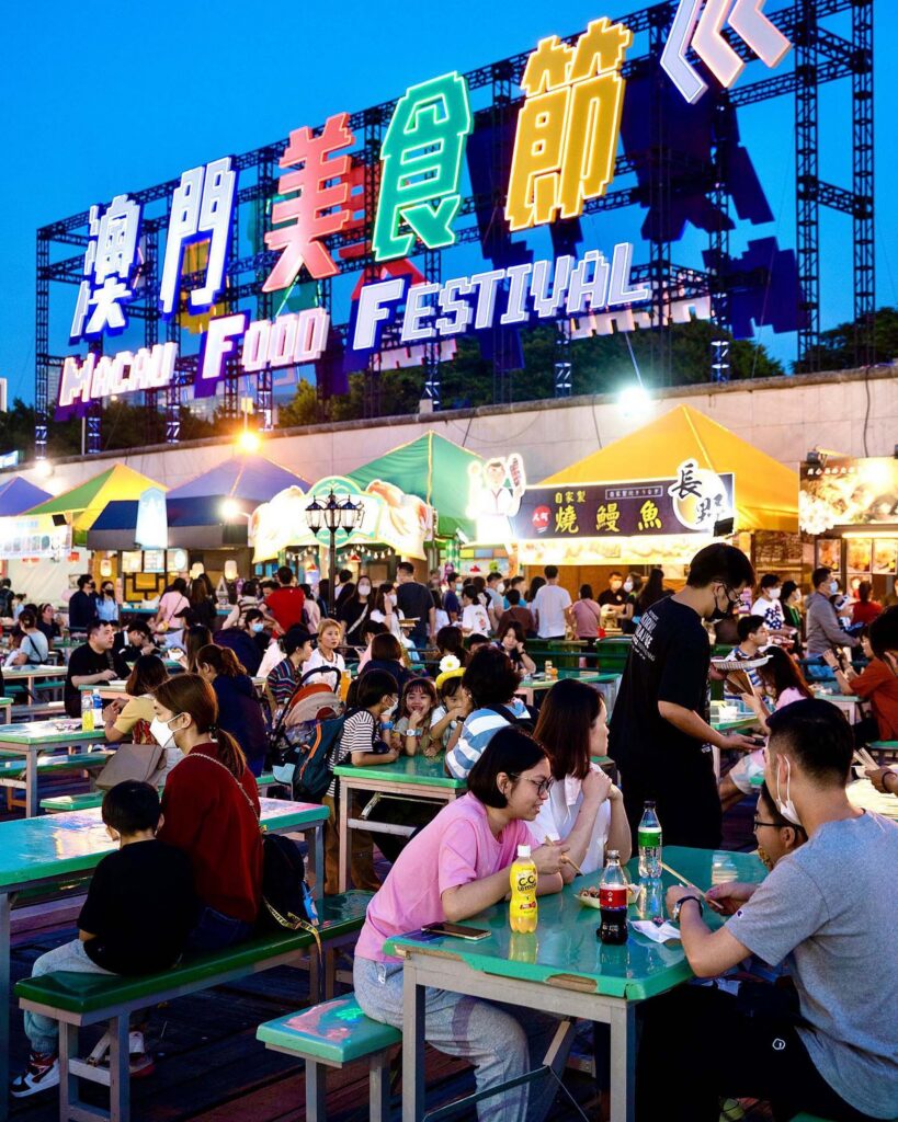Crowds eating at outdoor stalls at night under the illuminated Macau Food Festival sign