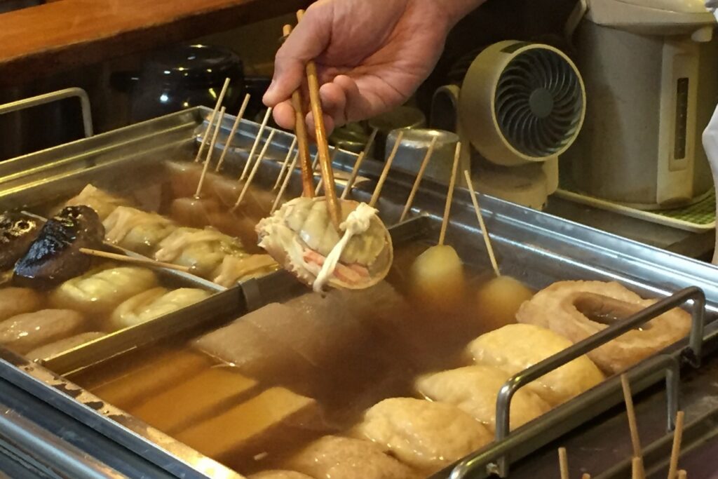 A hand lifting crab oden from a simmering oden pot with chopsticks in Kanazawa