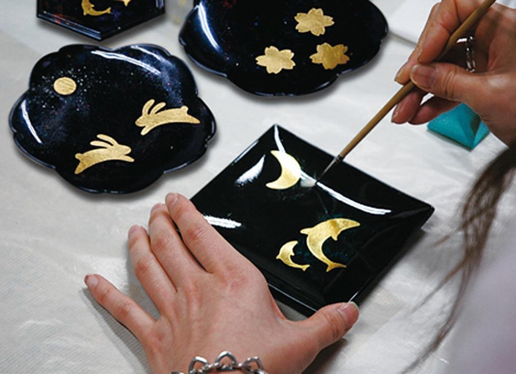 Hands applying gold-leaf motifs onto small black lacquer dishes with a brush