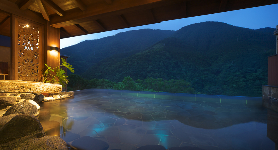 A private stone open-air bath at dusk with a carved wooden screen and a mountain view