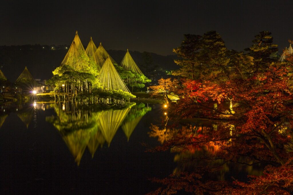 Kenroku-en Garden lit at night, glowing yukitsuri cones and autumn maples reflected in a pond