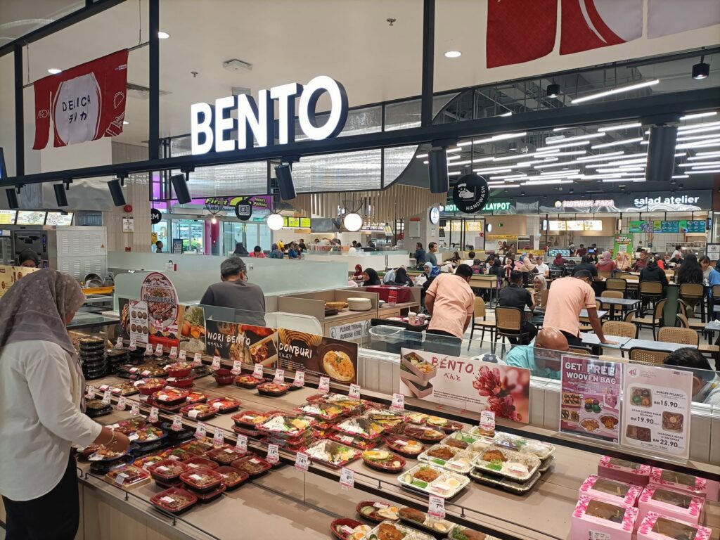 A Bento deli counter stacked with packed rice boxes, with food court seating behind